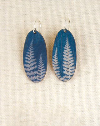 A pair of oval wooden drop earrings with a blue and white fern pattern, hanging from silver fishhook wires on a textured beige fabric background.