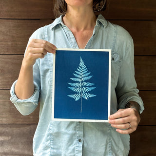 A person in a light blue shirt holds up a cyanotype art print featuring a white fern frond centered on a deep blue background, set against a dark wood plank wall.