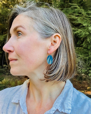 A woman with short gray-blonde hair is shown in profile outdoors, wearing a wood cyanotype earring with blue and white botanical designs. She is wearing a white button-up shirt, with a blurred green forest background.