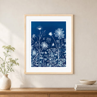 A framed botanical art print with white wildflower silhouettes on a deep blue background, displayed on a beige wall above a wooden console table with a vase and books.
