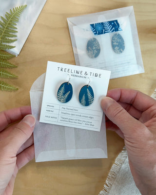 A person's hands hold a display card featuring a pair of oval cyanotype earrings with blue fern motifs on a white backing card. Another pair of earrings in a clear envelope sits on a light wood tabletop in the background.