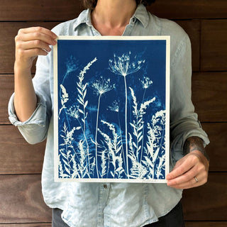 A person holds up a cyanotype art print featuring white wildflower silhouettes on a deep blue background, photographed against a dark wood plank wall.