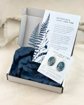 An open gift box on a beige fabric surface containing a pair of wood cyanotype blue earrings mounted on a branded card, along with a fern illustration and a tissue paper insert. The box lid has a printed card reading 'Botanicals, Light, and Time.