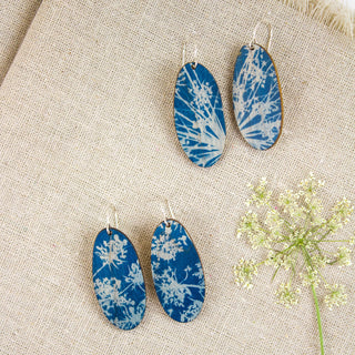 Three pairs of cyanotype oval drop earrings with blue and white floral patterns, displayed on a beige linen fabric background with a small white flower stem as styling.