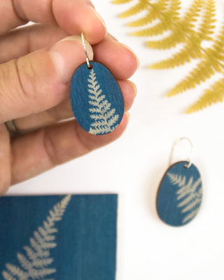 A hand holds an oval cyanotype blue wooden earring with a fern pattern, next to a matching wooden earring on a white background with a gold fern leaf prop.